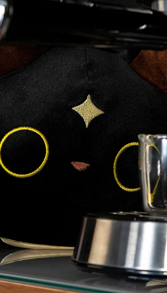 Cafemi black cat plush photographed beside coffee-making equipment